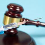 A person wearing surgical gloves holds a syringe. In the background a judge’s gavel.